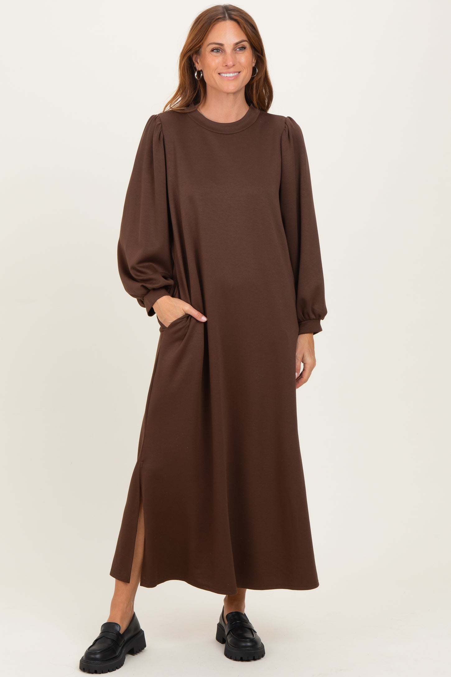 woman wearing a brown long sleeve maxi dress with puff sleeves and side pockets, standing and smiling