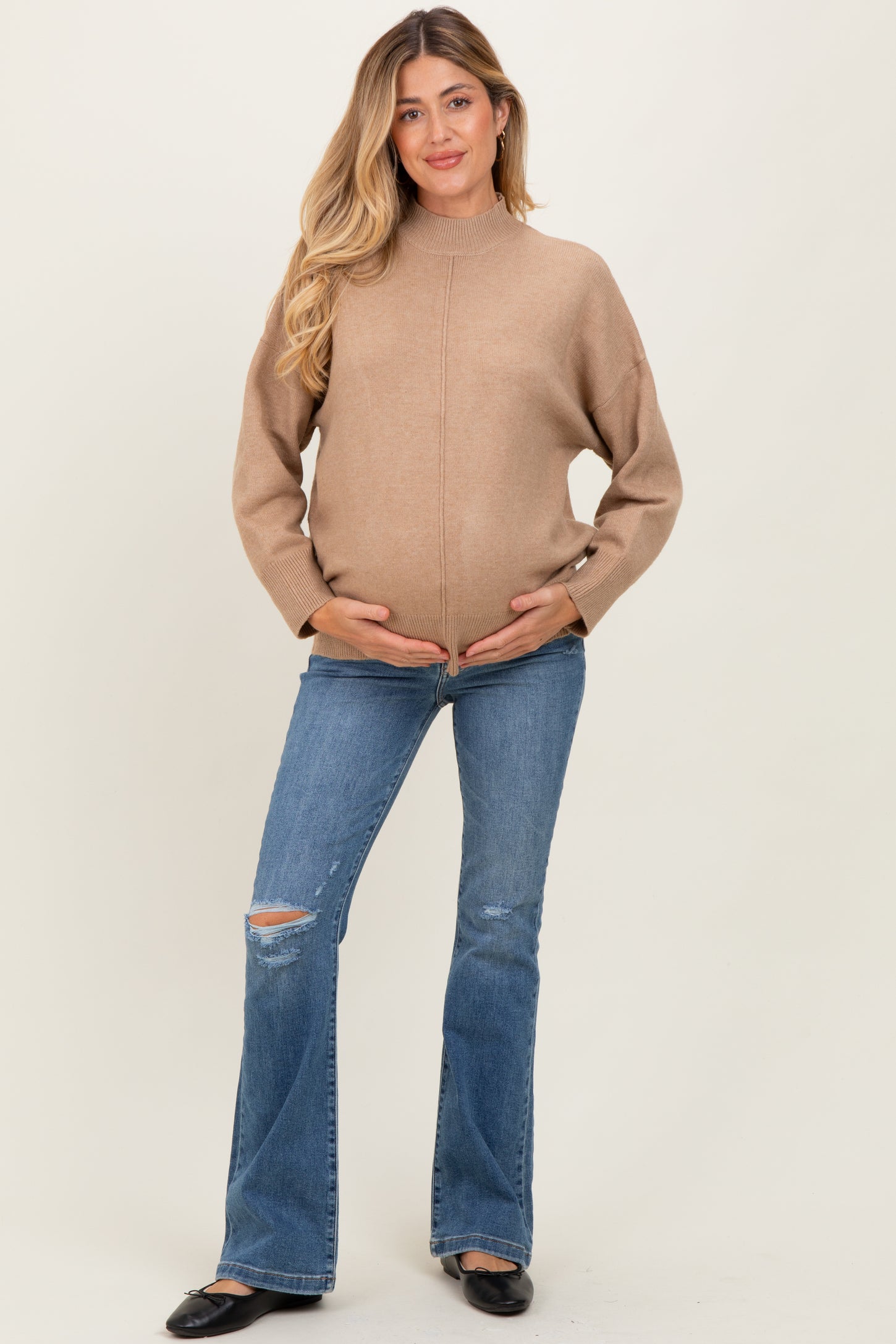 woman wearing a beige maternity sweater and blue distressed flared jeans, holding her baby bump