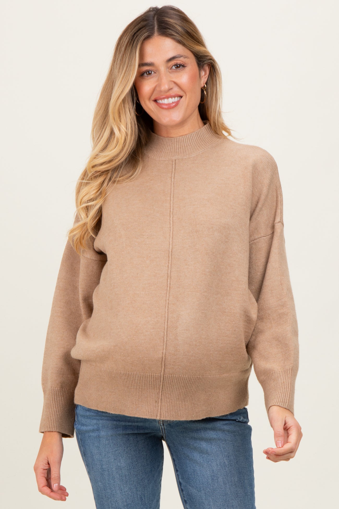 woman wearing a tan maternity sweater with a mock neck and relaxed fit, smiling while holding her baby bump