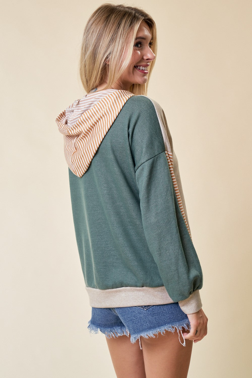 woman wearing a green and beige color block hoodie with a striped hood and denim shorts