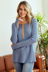 woman wearing a blue long sleeve henley top with orange button details and relaxed fit, smiling indoors