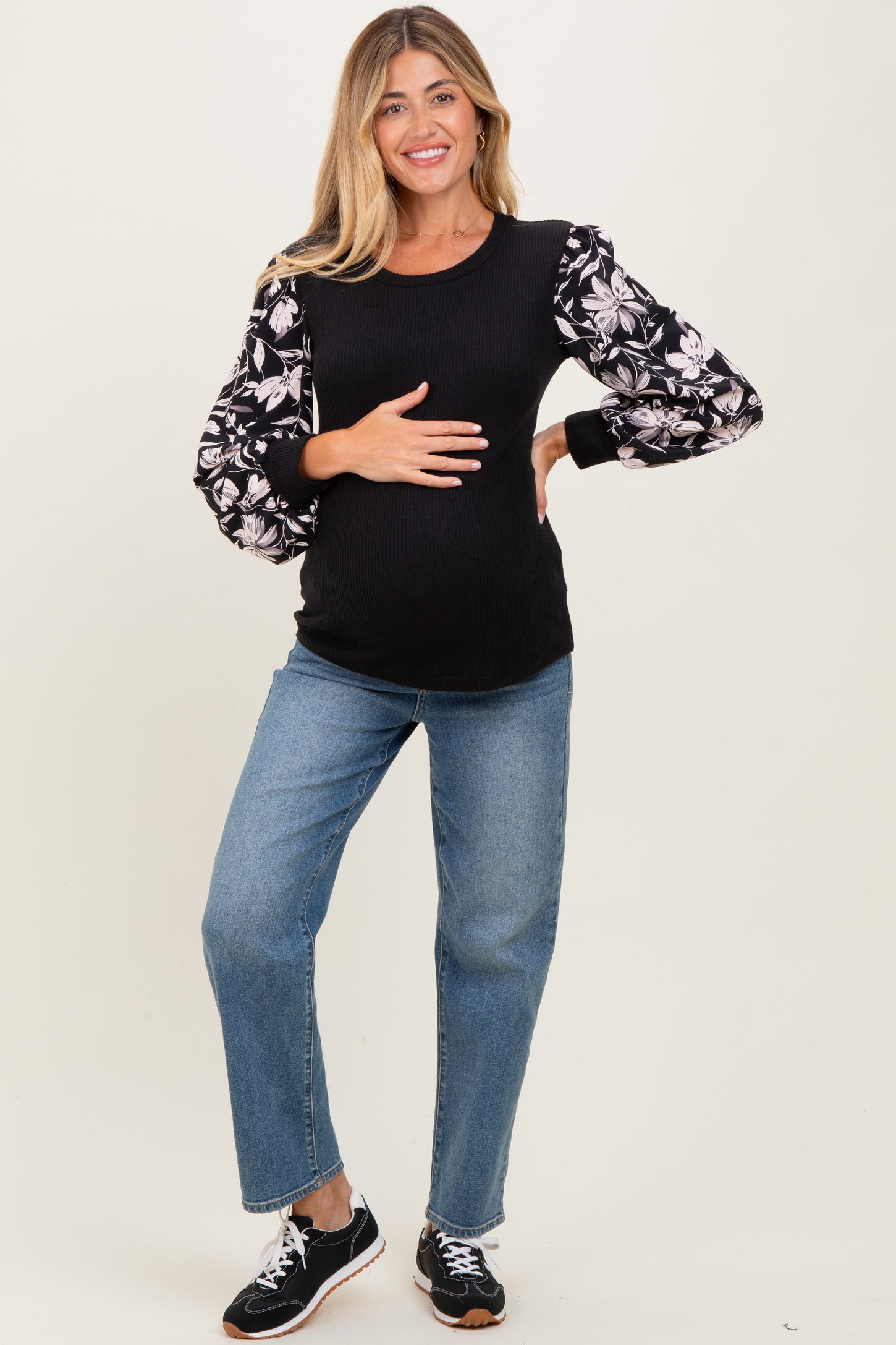 woman wearing a black maternity top with floral puff sleeves and holding her baby bump, paired with light wash jeans