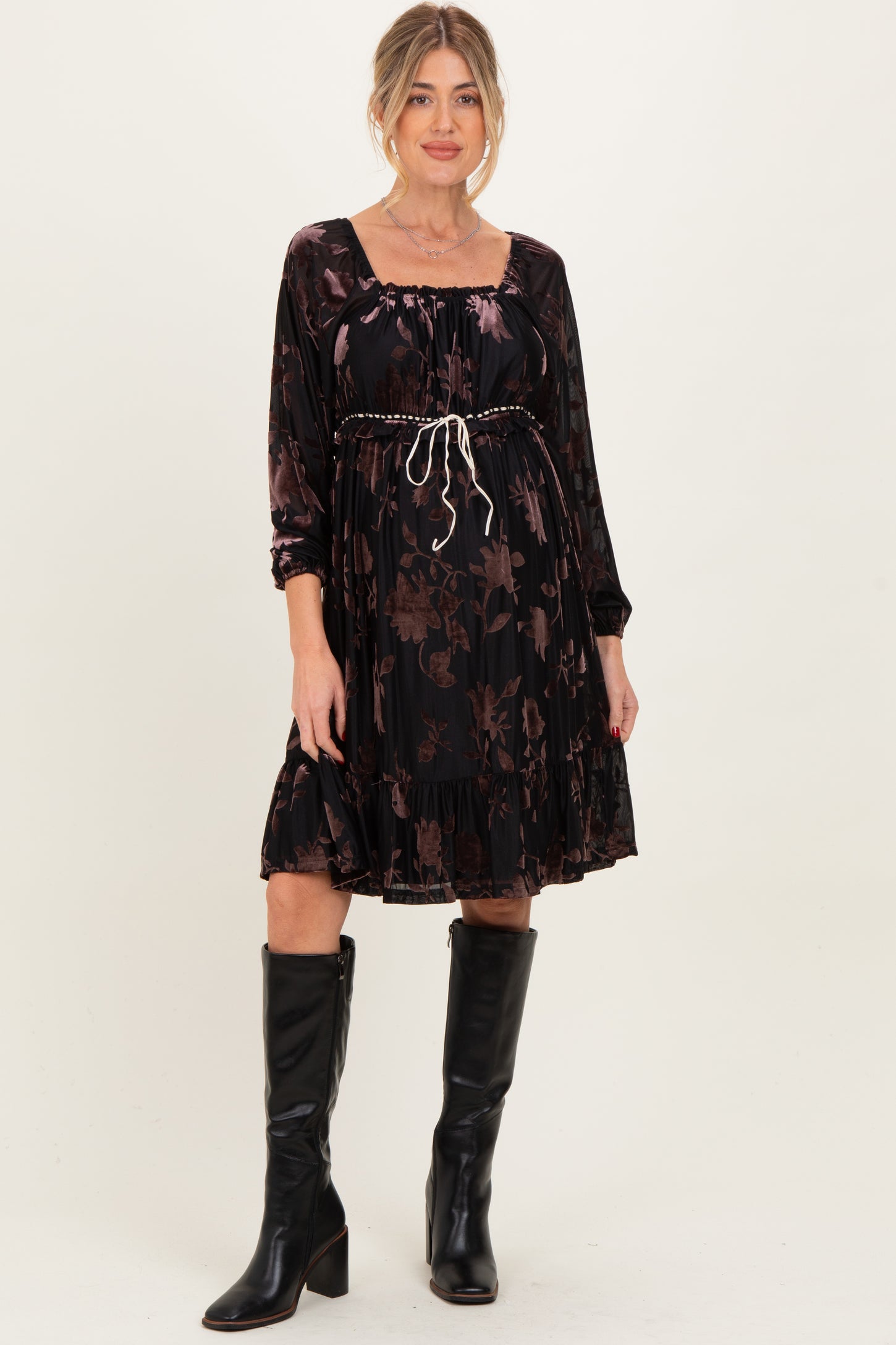 woman wearing a black floral maternity dress with long sleeves and a cinched waist, paired with knee-high black boots