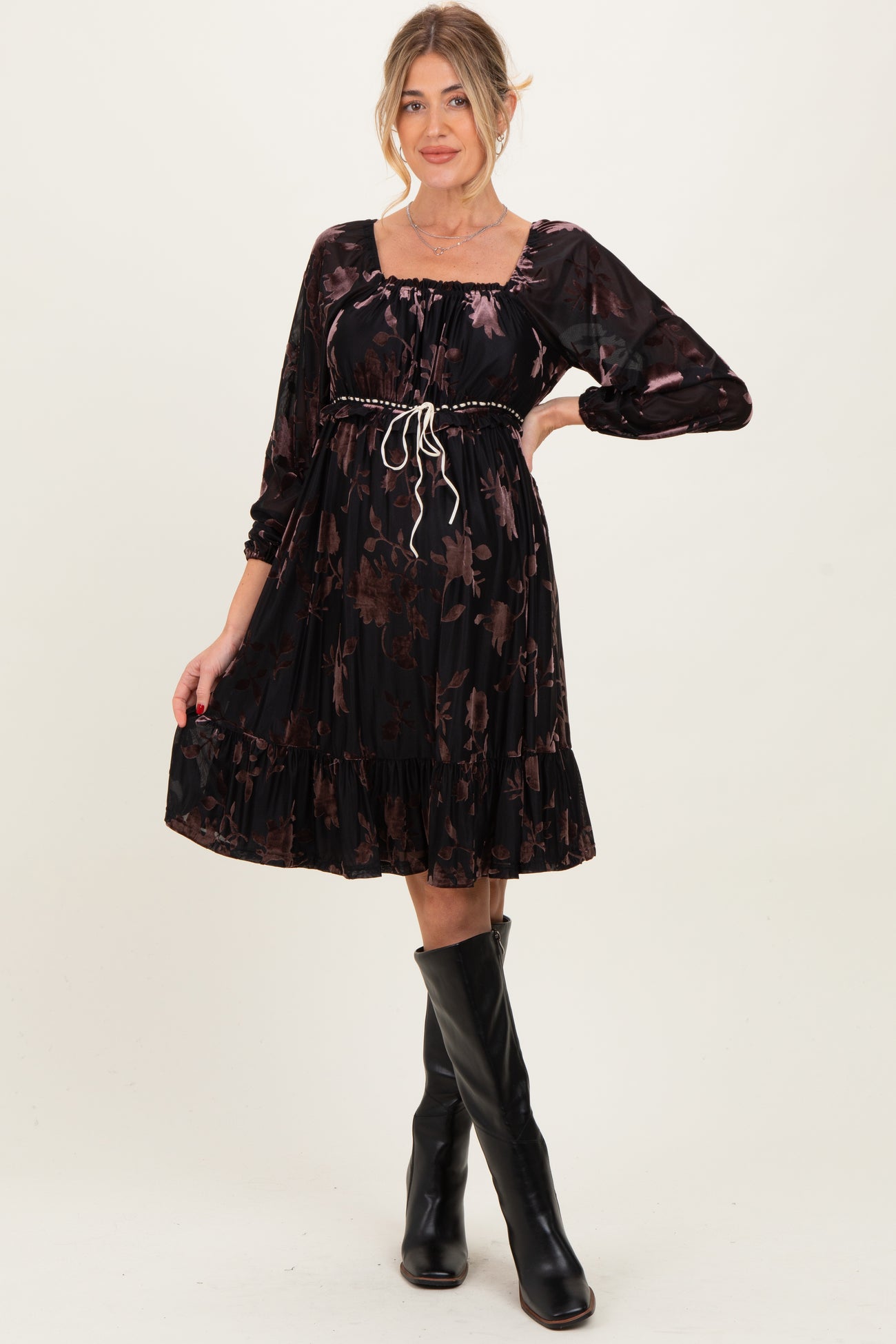 woman wearing a black floral maternity dress with long sleeves and a cinched waist, styled with knee-high black boots