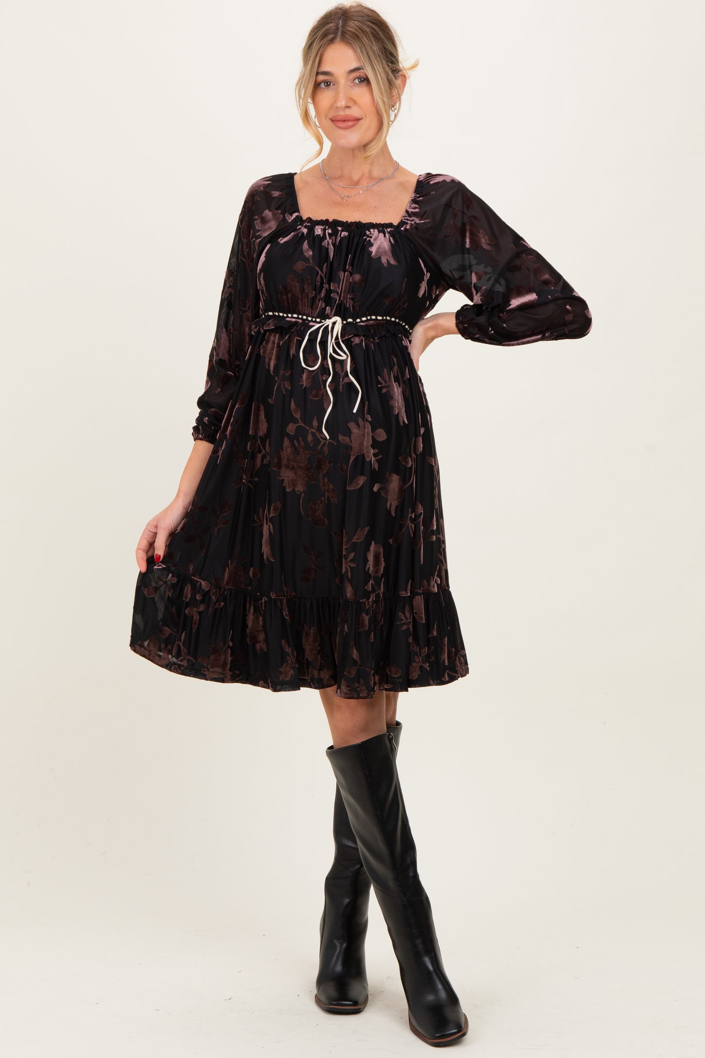 woman wearing a black floral maternity dress with long sleeves and a cinched waist, styled with knee-high black boots