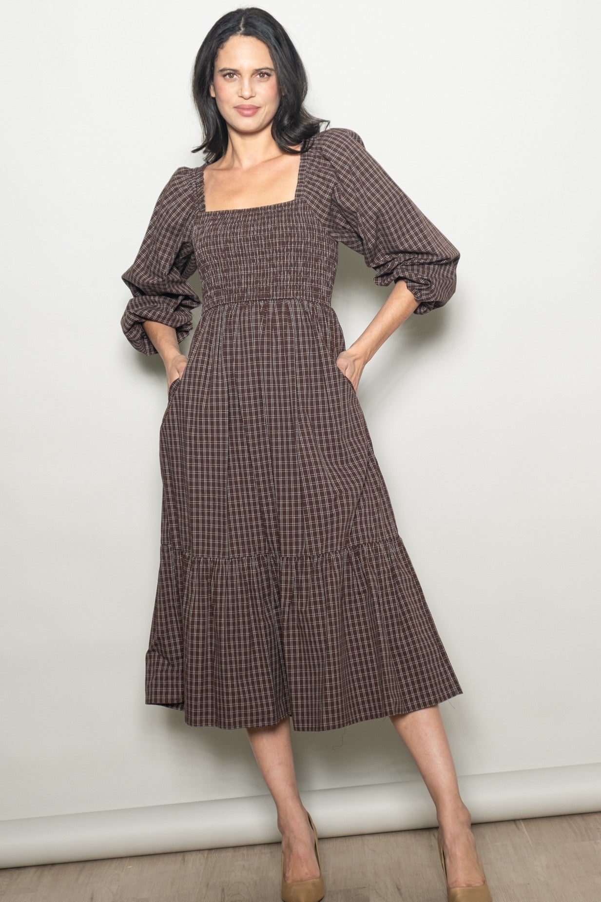 woman wearing a brown checkered midi dress with a square neckline and puff sleeves, standing with hands in pockets
