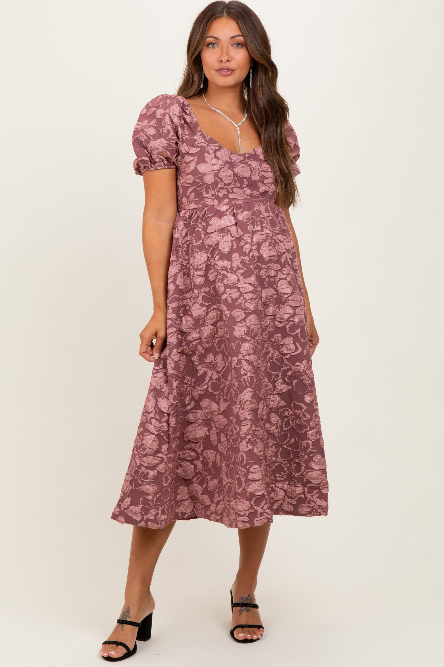 woman wearing a mauve floral maternity dress with puff sleeves and a flowy skirt, posing with a relaxed expression