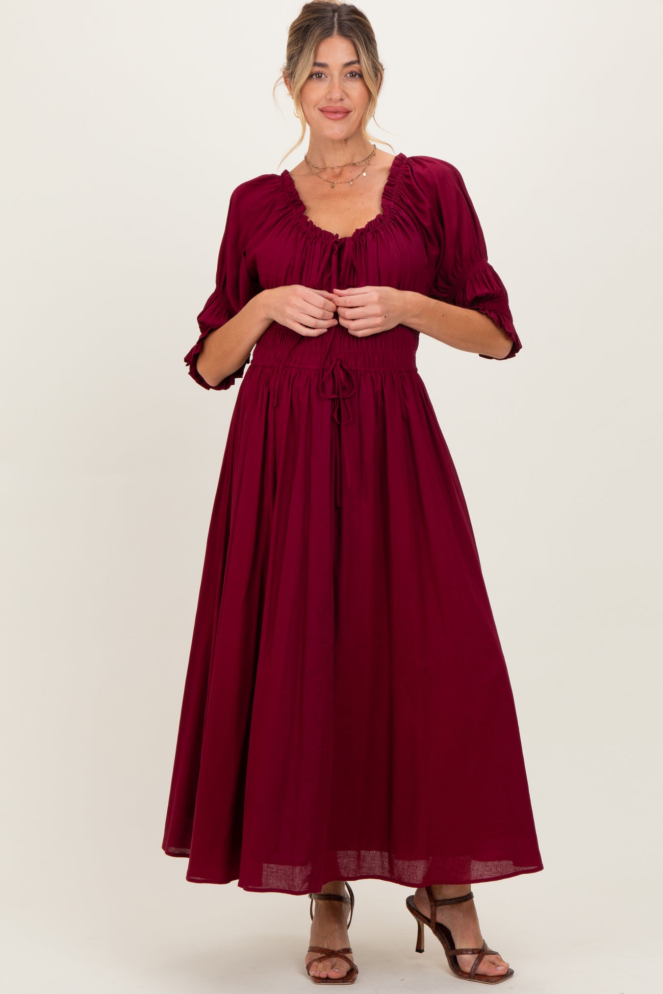 woman wearing a burgundy maternity maxi dress with puff sleeves and a drawstring waist, holding the fabric at the front