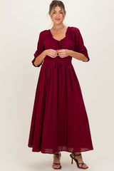 woman wearing a burgundy maternity maxi dress with puff sleeves and a drawstring waist, holding the fabric at the front