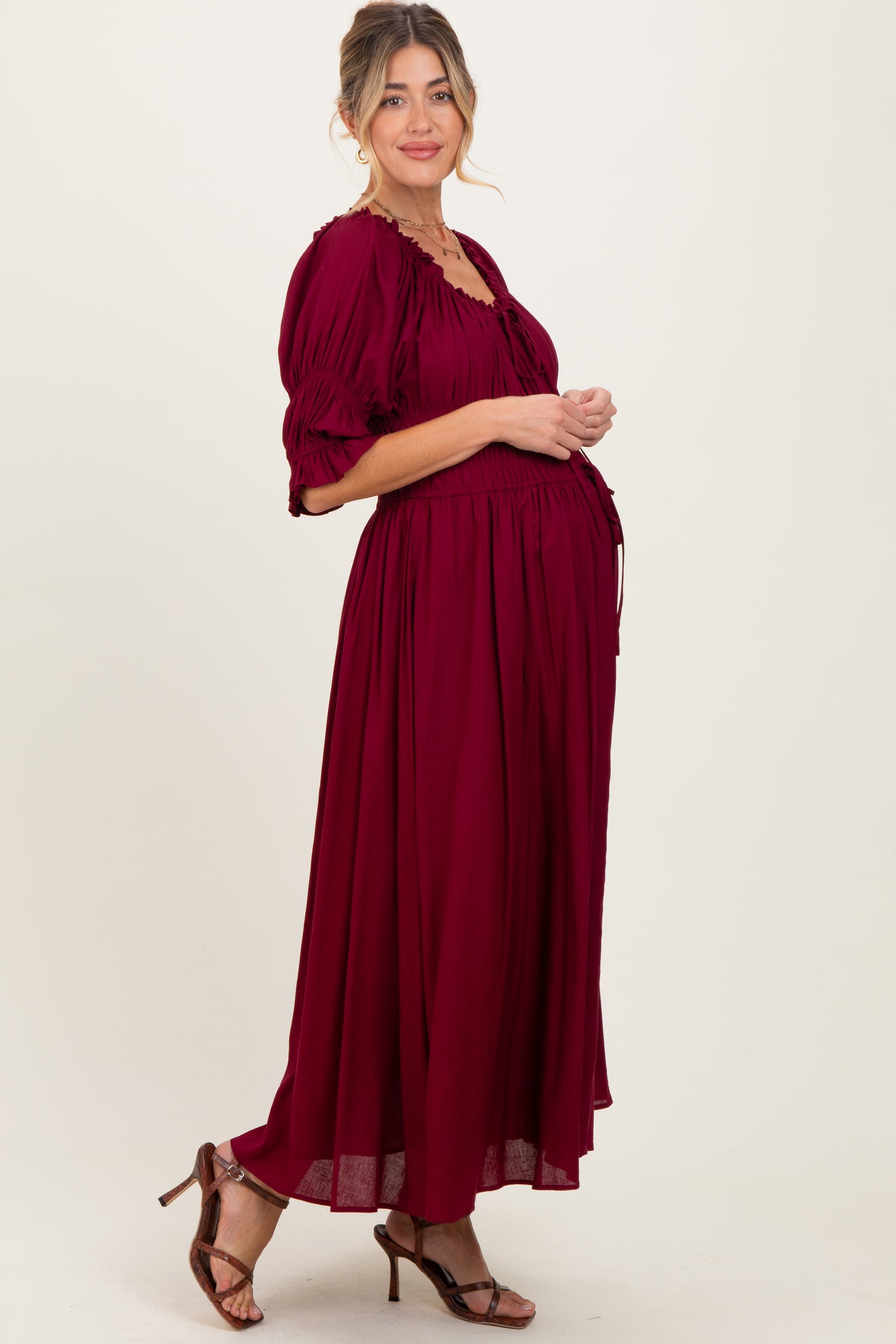 woman wearing a burgundy maternity maxi dress with puff sleeves and a fitted bodice, showcasing her baby bump
