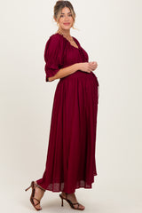 woman wearing a burgundy maternity maxi dress with puff sleeves and a fitted bodice, showcasing her baby bump
