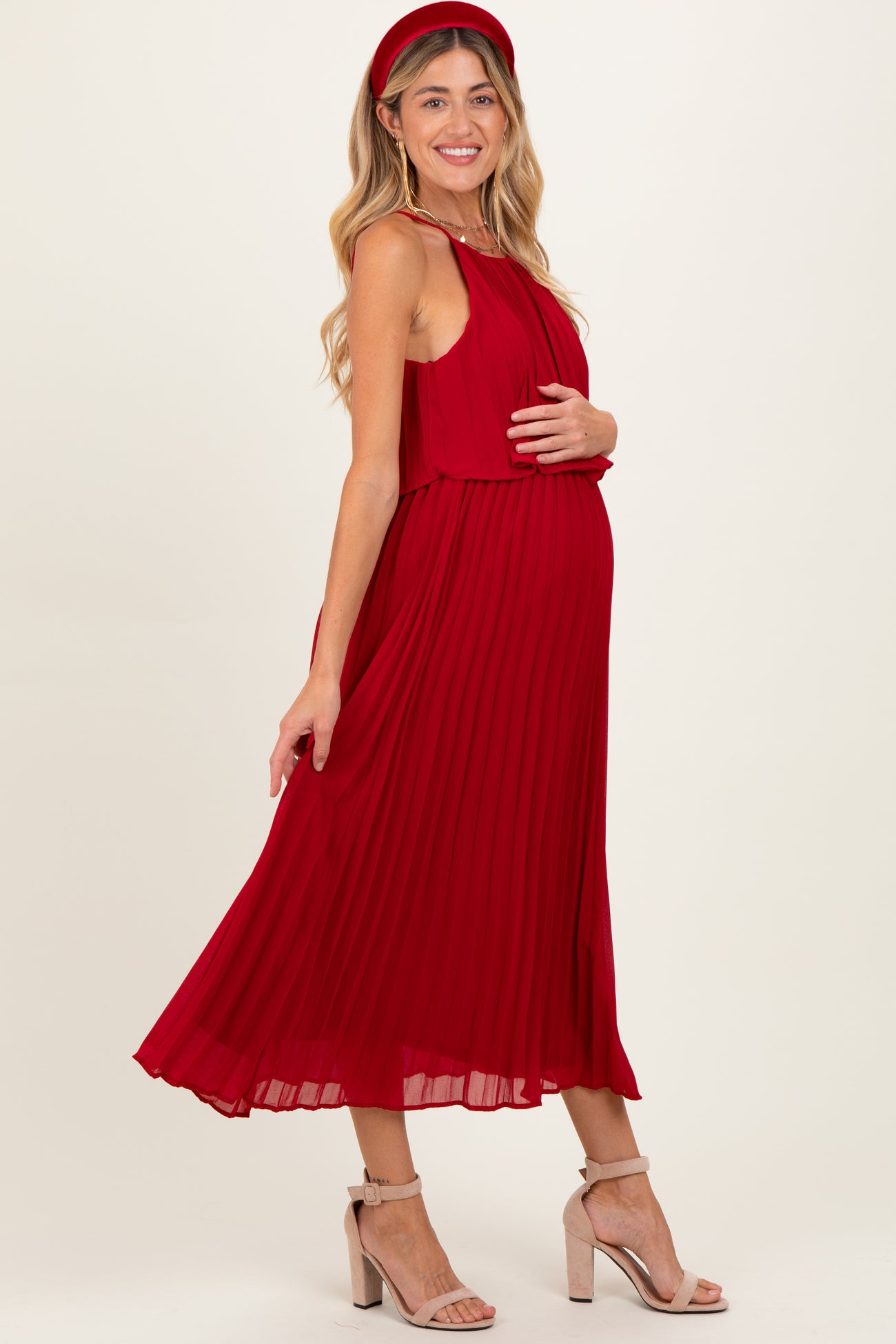 woman wearing a red pleated maternity dress, showing off her baby bump, paired with a matching headband and heels