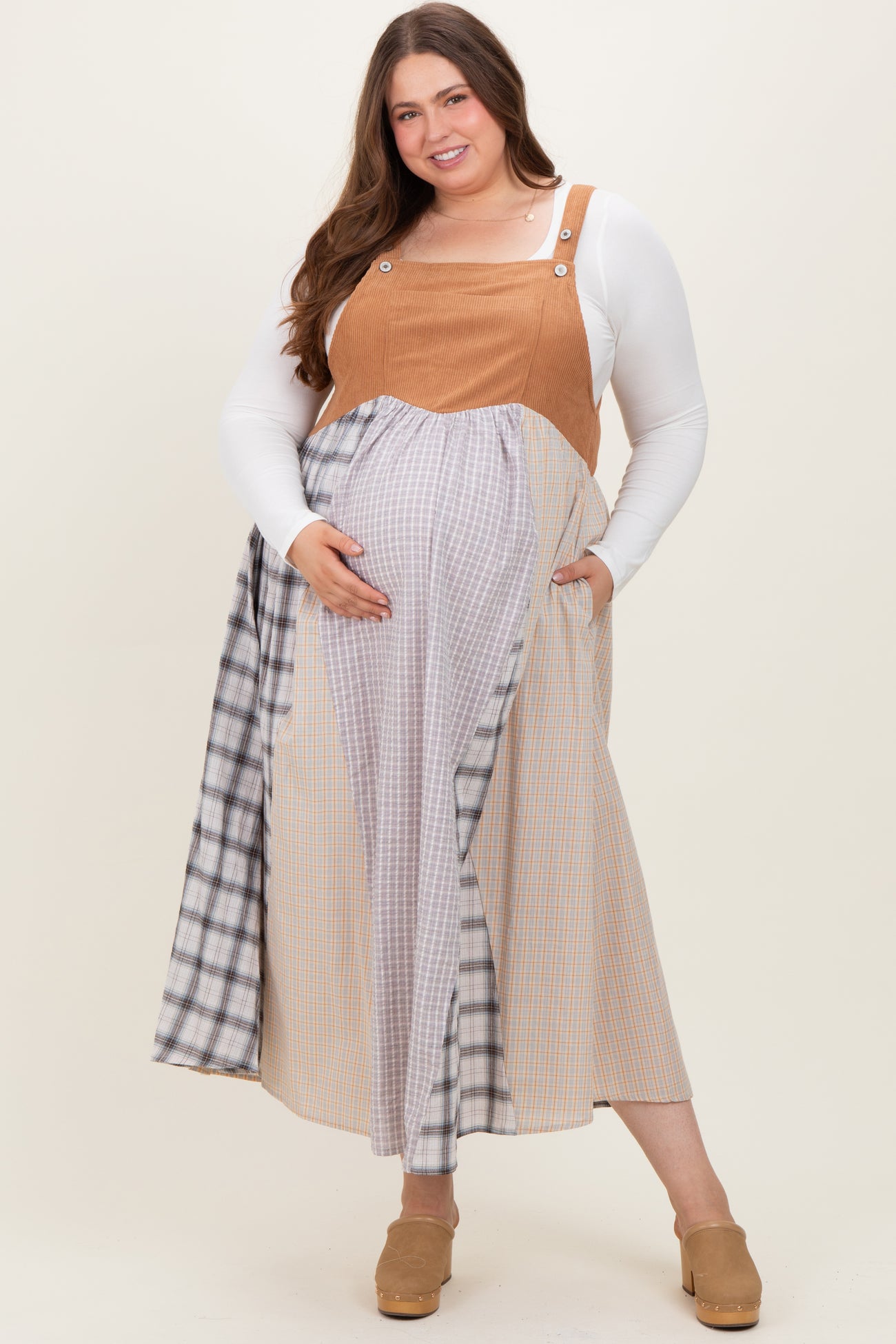 woman wearing a tan maternity plus overall dress with plaid detailing, showing her baby bump and smiling