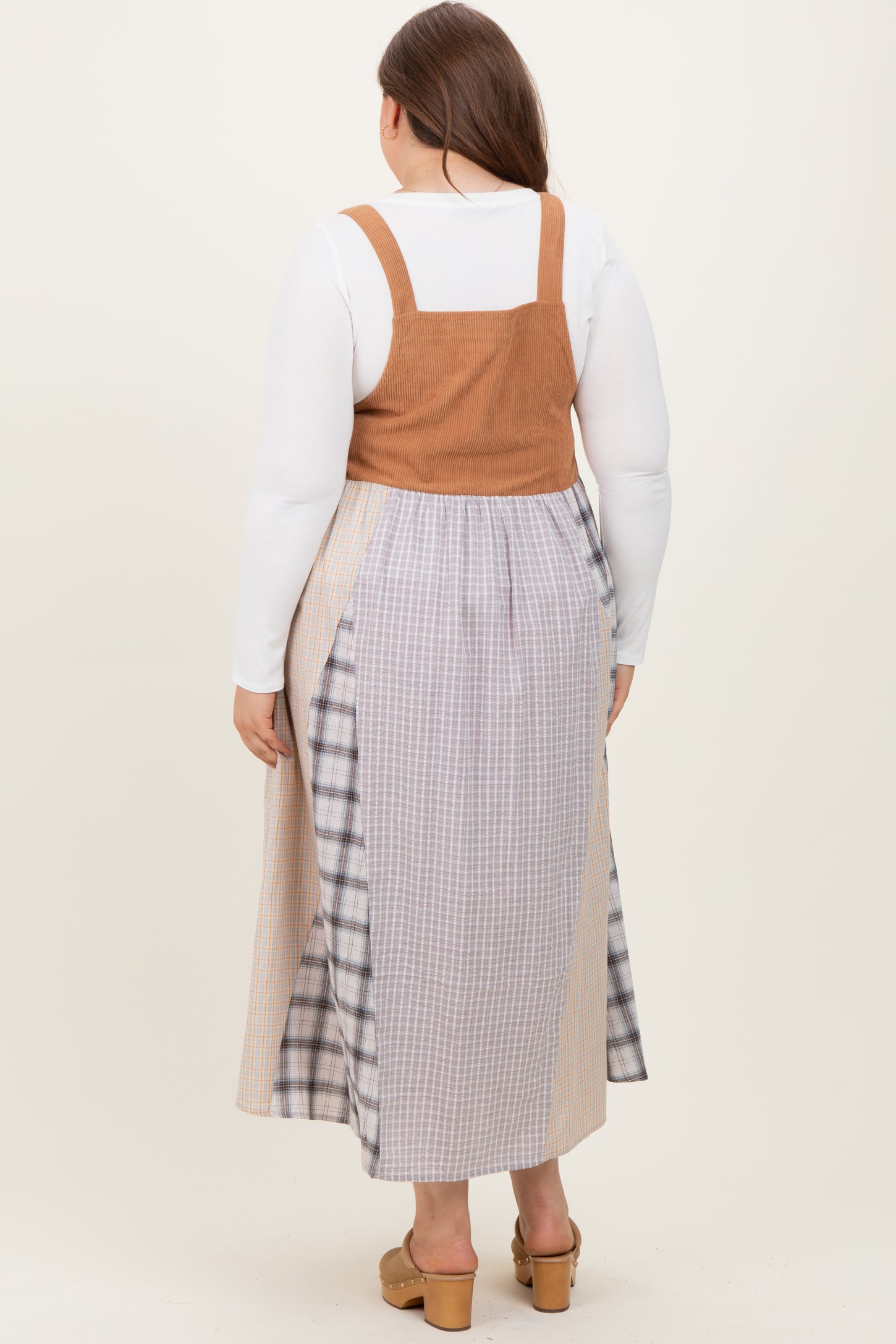 woman wearing a maternity plus tan corduroy overall dress with a long plaid skirt, viewed from the back
