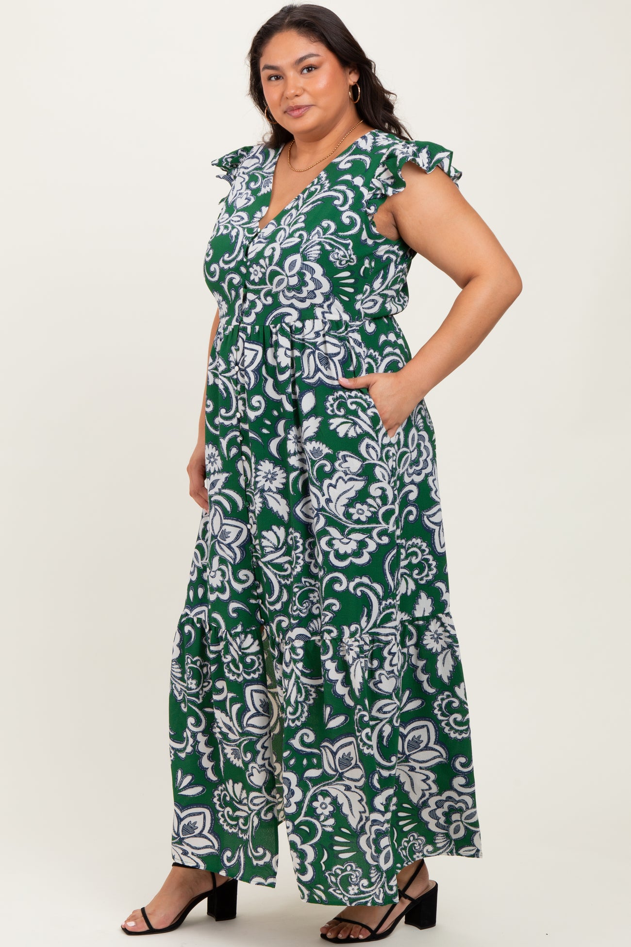 woman wearing a green and white floral plus-size maxi dress with short flutter sleeves and a tie waist, posed with hands in pockets