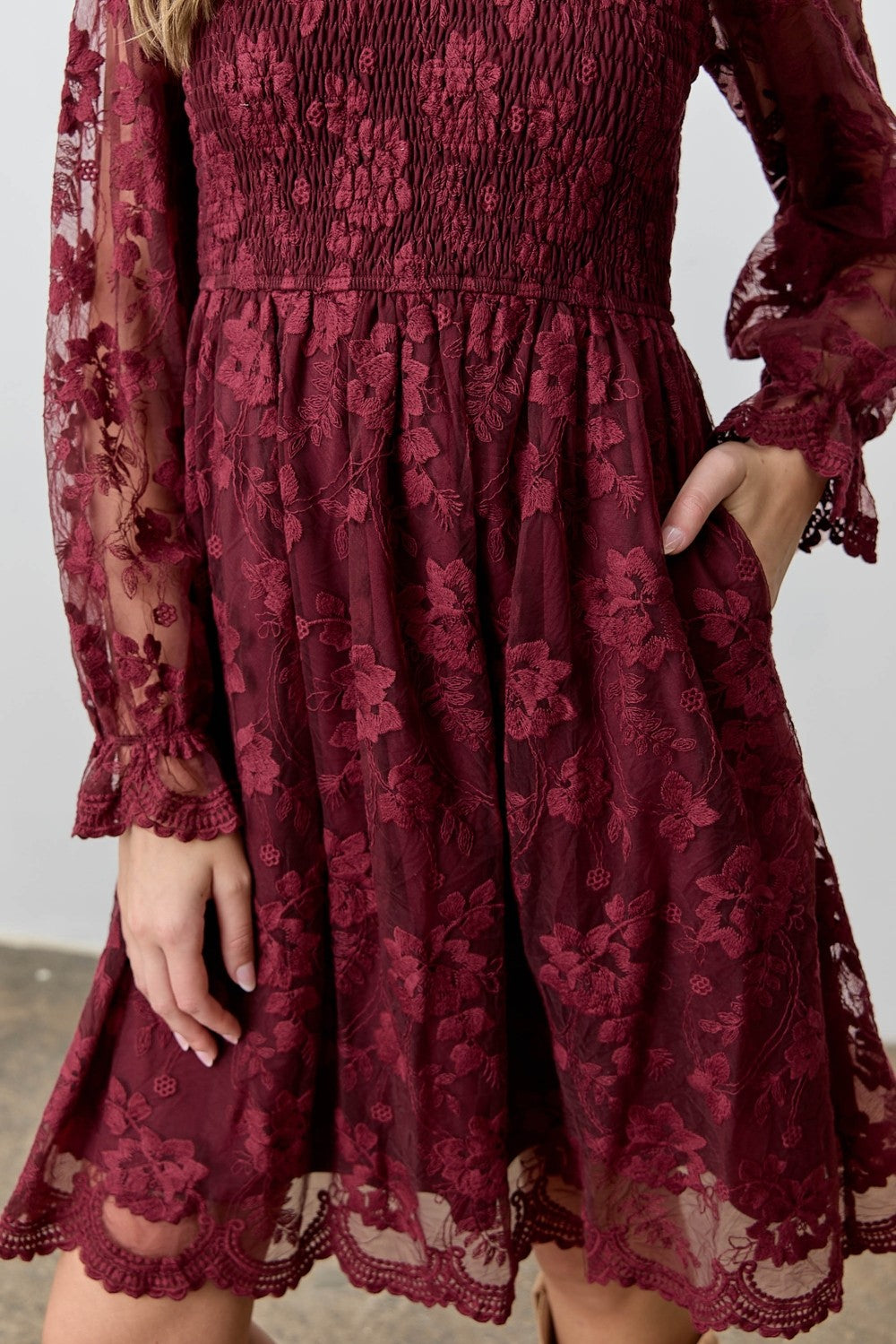 woman wearing a burgundy lace dress with smocked bodice and long sheer sleeves, featuring floral embroidery and pockets