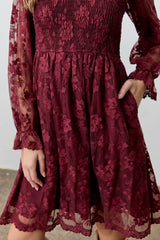 woman wearing a burgundy lace dress with smocked bodice and long sheer sleeves, featuring floral embroidery and pockets