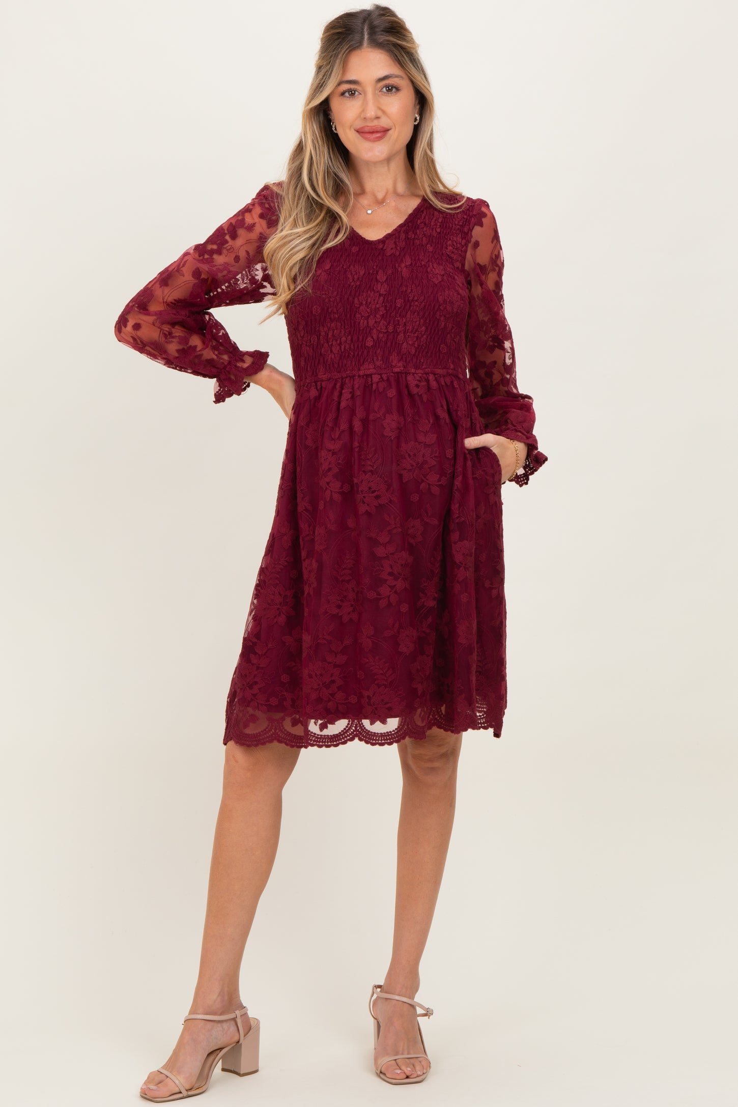 woman wearing a burgundy lace maternity dress with long sheer sleeves and pockets, posing with a slight smile