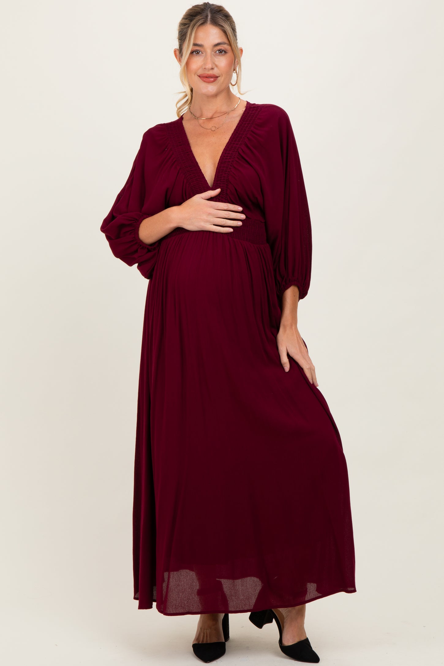 woman wearing a burgundy maternity maxi dress with a deep v-neck and flowing sleeves, holding her baby bump