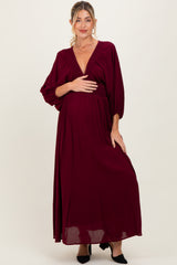 woman wearing a burgundy maternity maxi dress with a deep v-neck and flowing sleeves, holding her baby bump