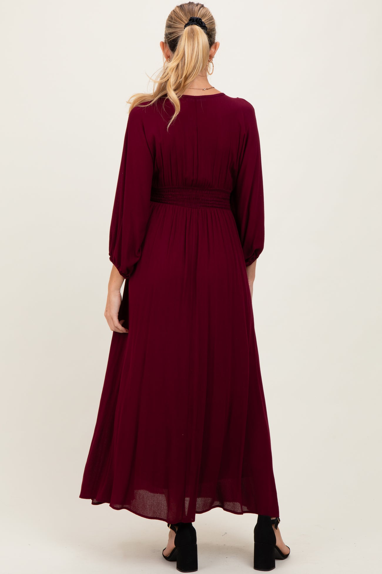 woman wearing a burgundy maternity maxi dress with a smocked waist and long balloon sleeves, showing the back view