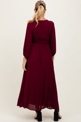 woman wearing a burgundy maternity maxi dress with a smocked waist and long balloon sleeves, showing the back view