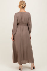 woman wearing a long taupe maternity dress with a smocked waist and long sleeves, viewed from the back