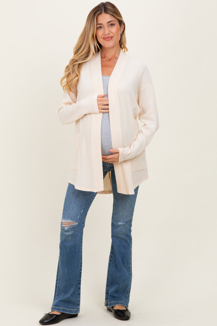 woman wearing a beige long cardigan over a tank top and holding her baby bump while standing in distressed blue jeans