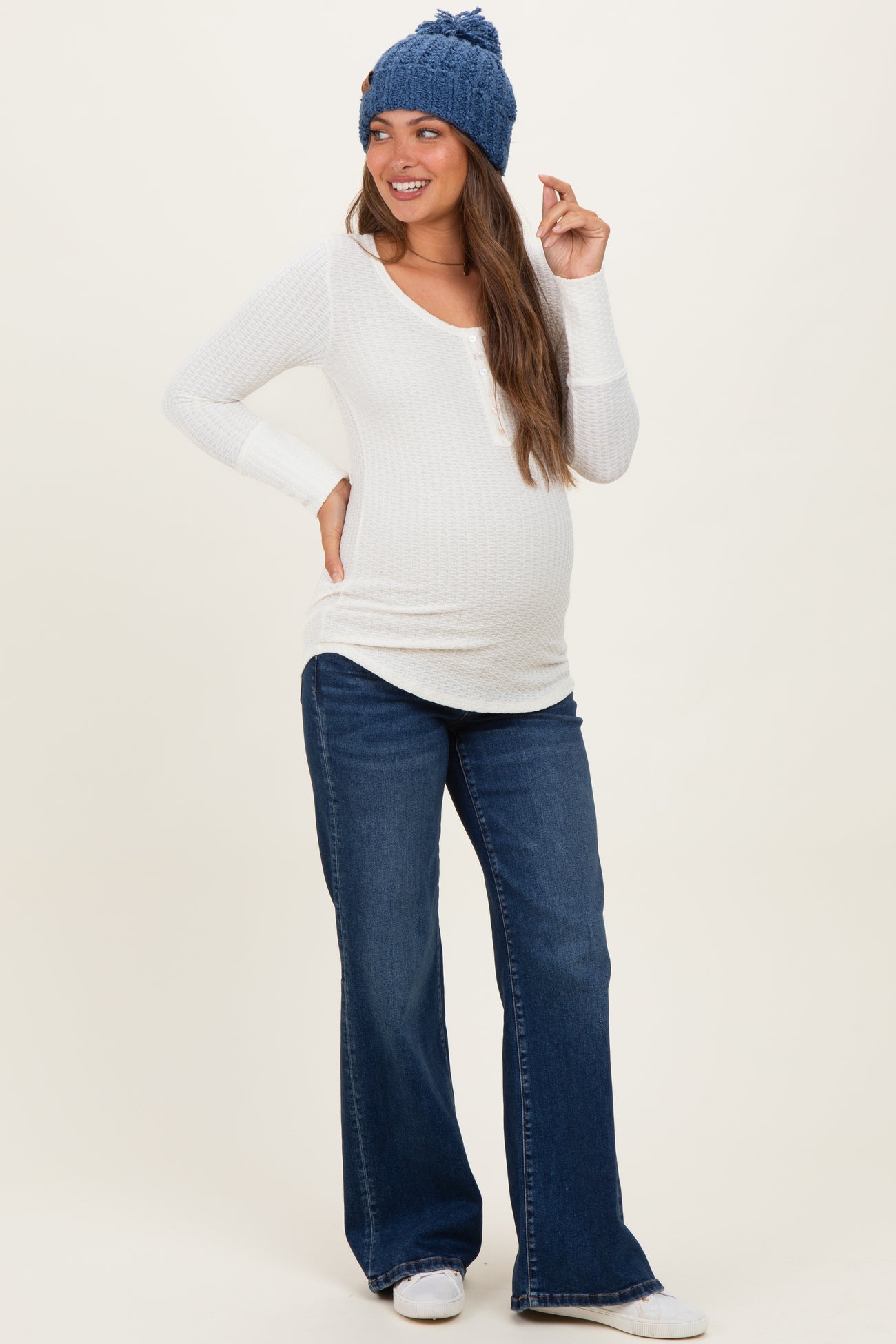 woman wearing a white long sleeve maternity top and blue flared jeans, showing her baby bump and smiling