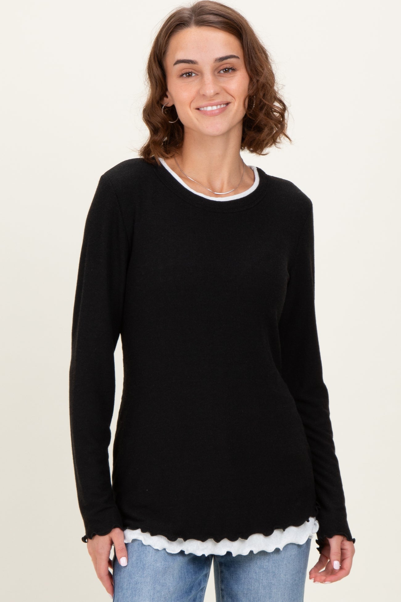 woman wearing a black long sleeve top with a white scalloped hem over a white shirt, smiling against a light background