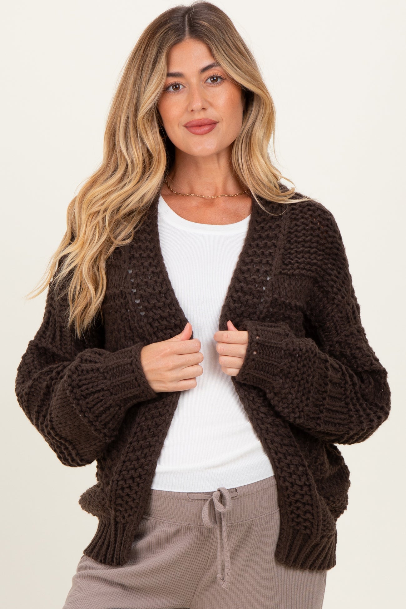 woman wearing a dark brown chunky knit maternity cardigan over a white top and gray pants, smiling at the camera