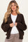 woman wearing a dark brown chunky knit maternity cardigan over a white top and gray pants, smiling at the camera