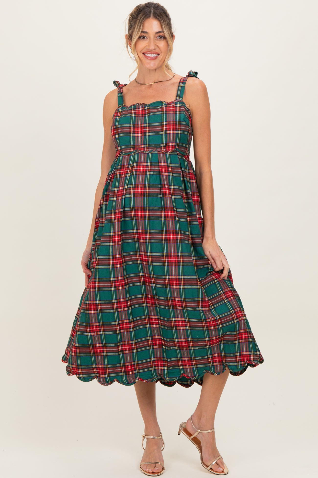 woman wearing a green and red plaid maternity dress with tie straps and a scalloped hem, showcasing her baby bump