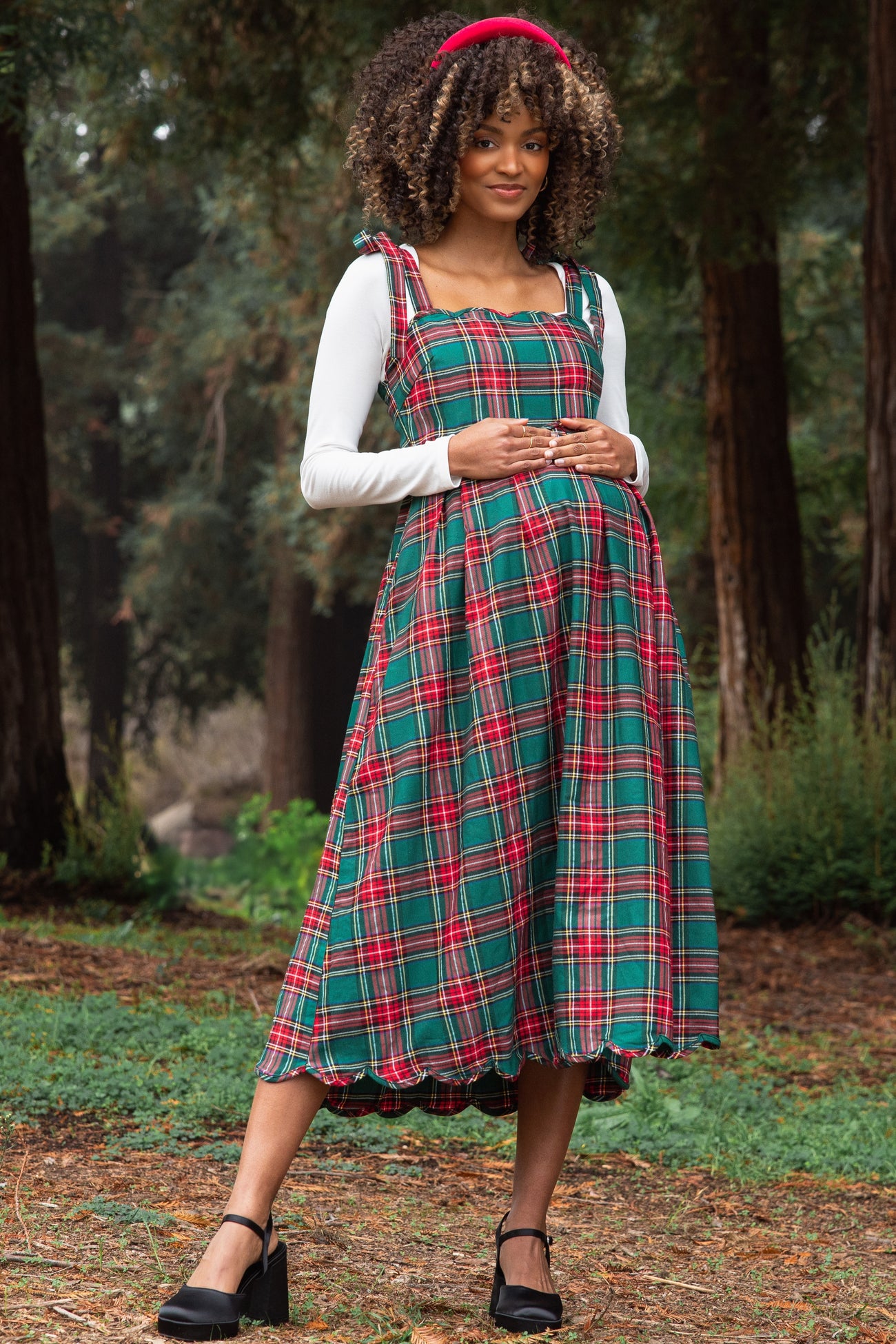 woman wearing a green and red plaid maternity dress with a white long sleeve top, holding her baby bump in an outdoor setting