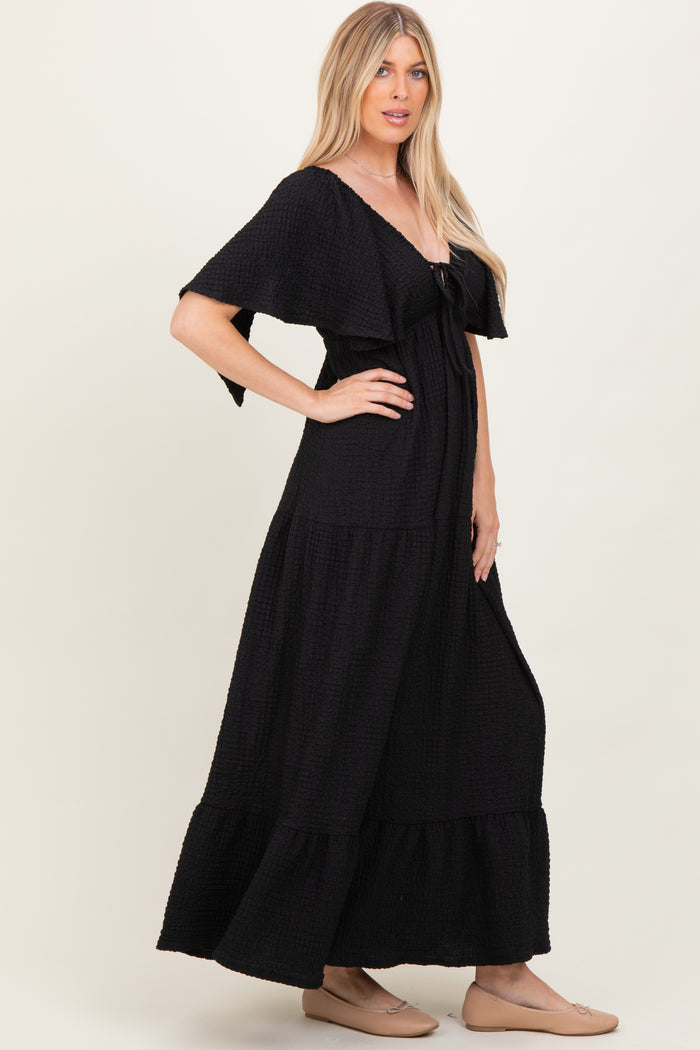 woman wearing a black maxi dress with a tiered design and flutter sleeves, standing in profile against a neutral background