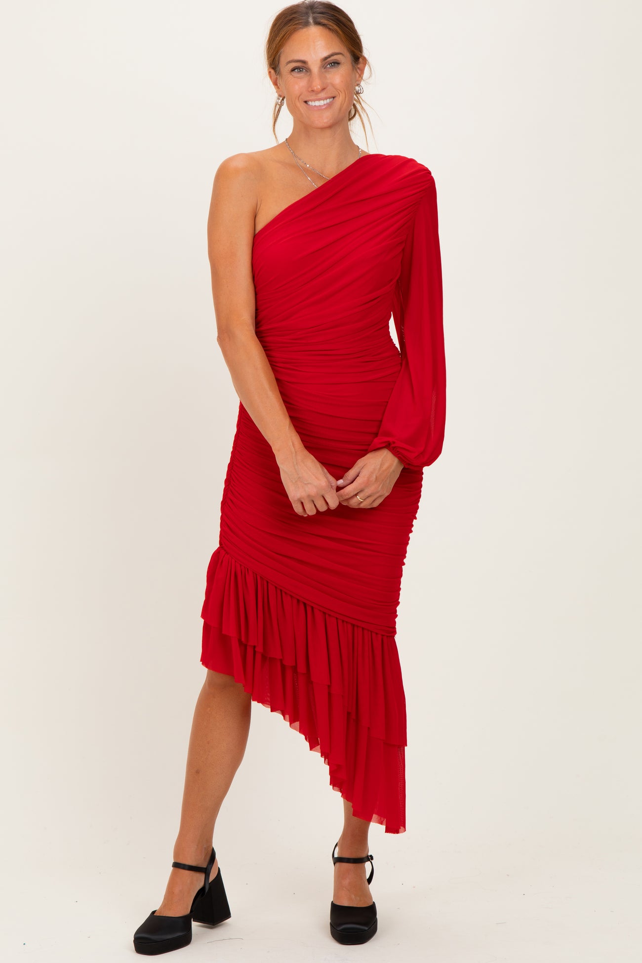 woman wearing a red one-shoulder dress with ruched detailing and a layered ruffle hem, standing in a relaxed pose