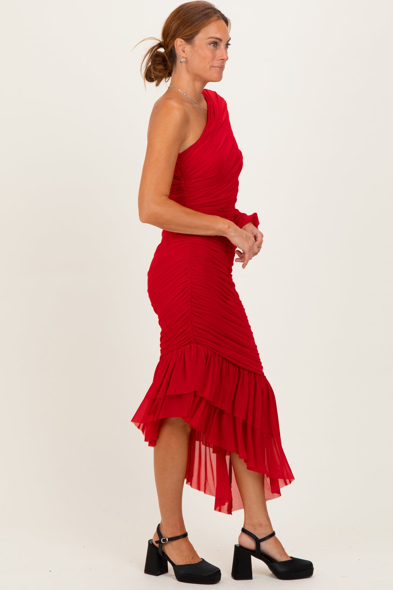 woman wearing a red one-shoulder fitted dress with ruched details and a ruffled hem, paired with black heeled sandals