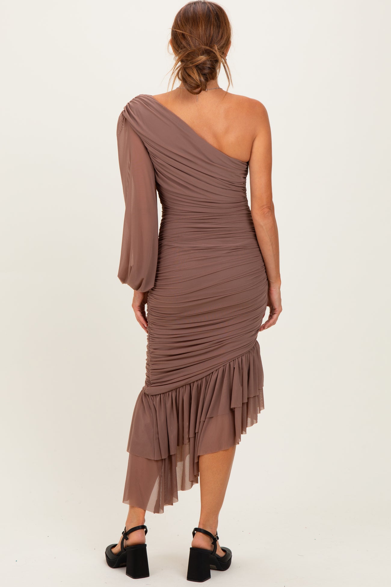 woman wearing a taupe one-shoulder ruched dress with a ruffled hem and long sleeve details, view from the back