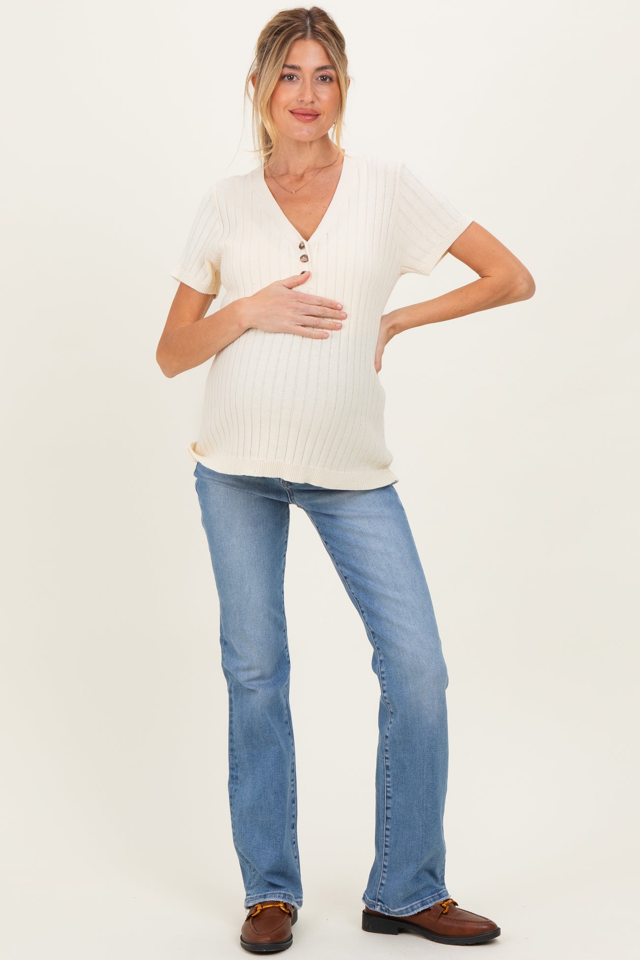 woman wearing a cream ribbed maternity top with buttons and blue flared jeans, gently holding her baby bump