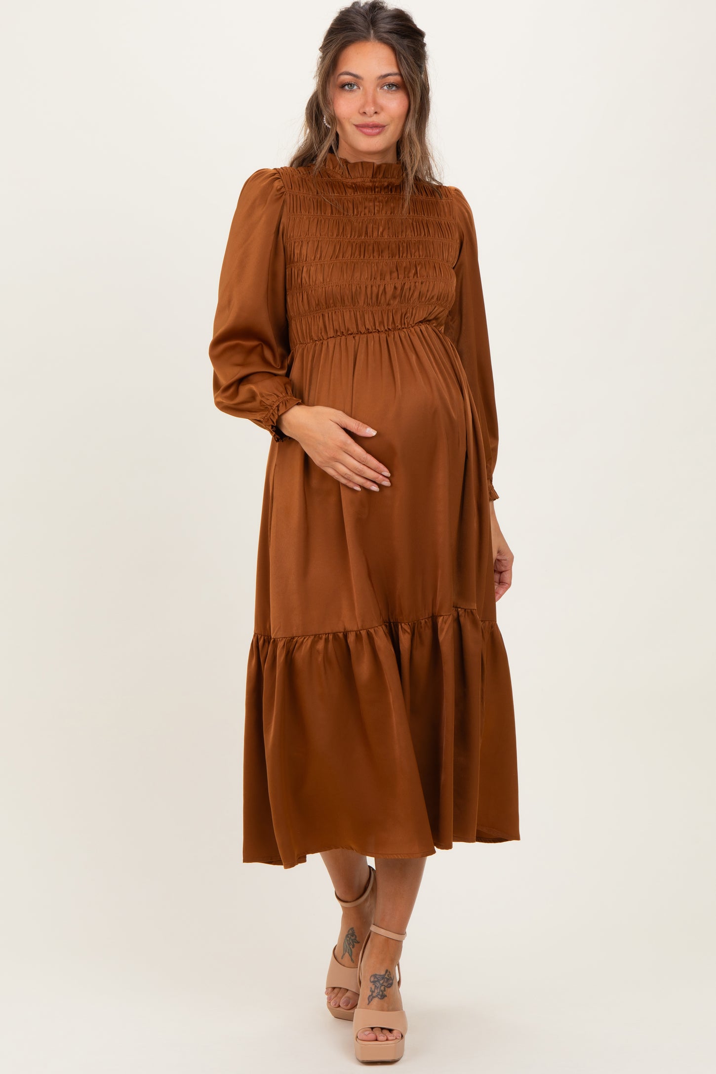 woman wearing a brown maternity dress with long sleeves and a smocked bodice, gently cradling her baby bump