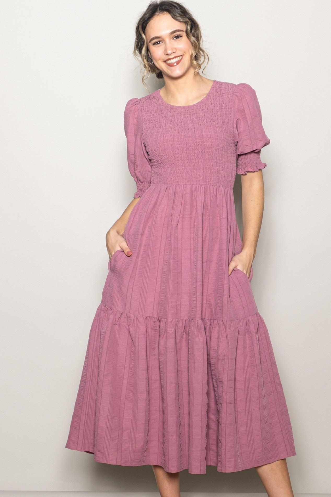 woman wearing a mauve puff sleeve maxi dress with a smocked bodice and tiered skirt, posing with hands in pockets
