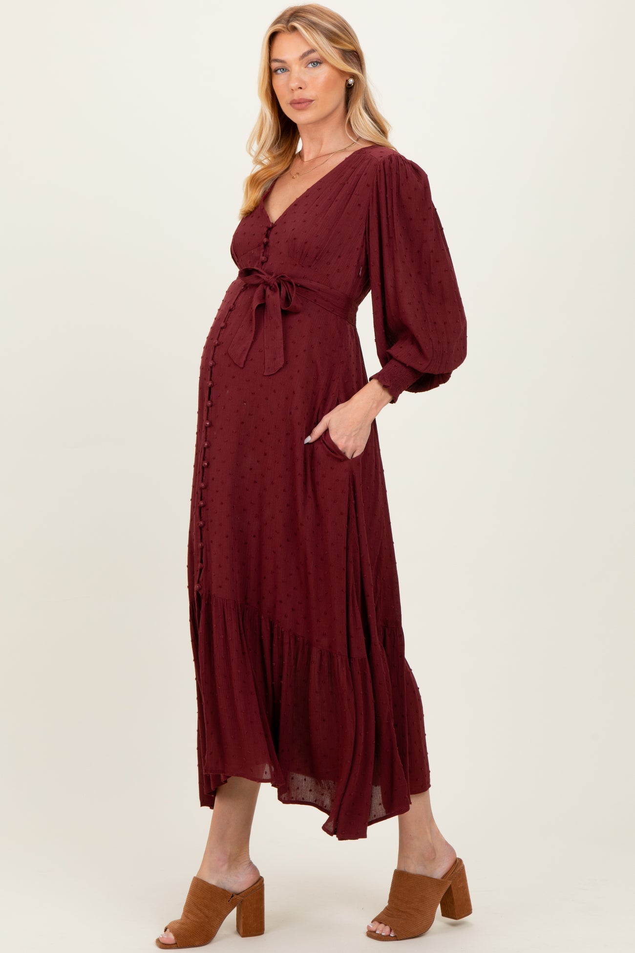 woman wearing a burgundy maternity dress with a tie waist, long sleeves, and a tiered skirt, displaying her baby bump