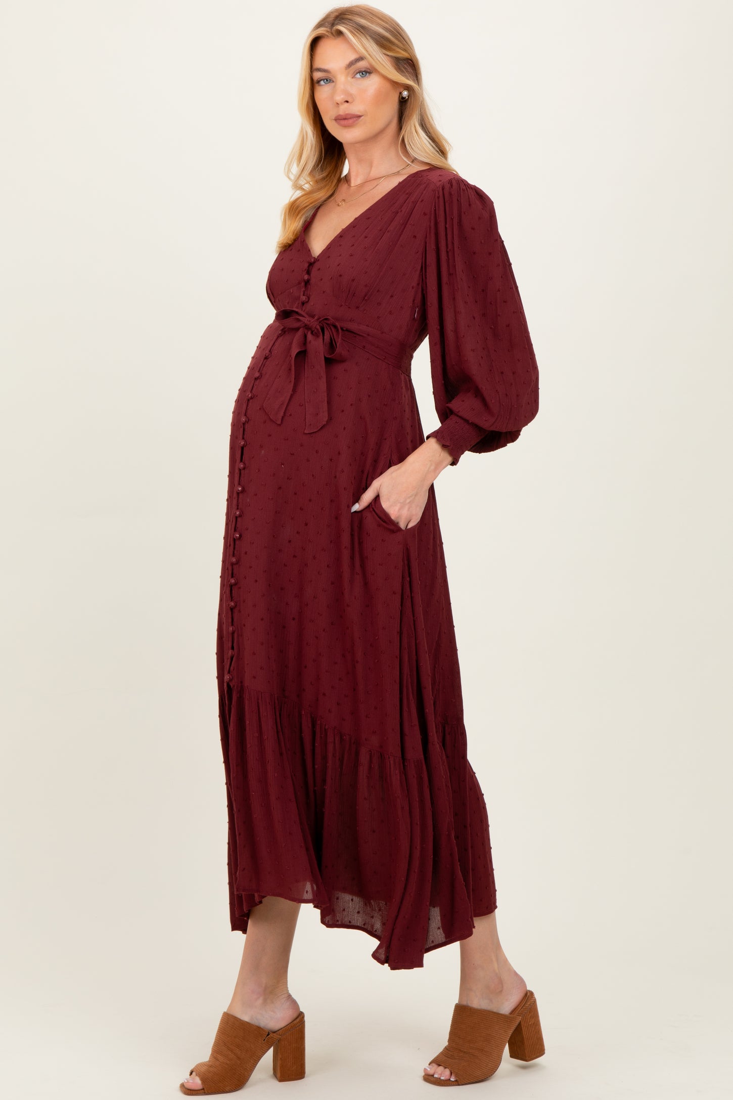 woman wearing a burgundy maternity dress with a tie waist, long sleeves, and a tiered skirt, displaying her baby bump