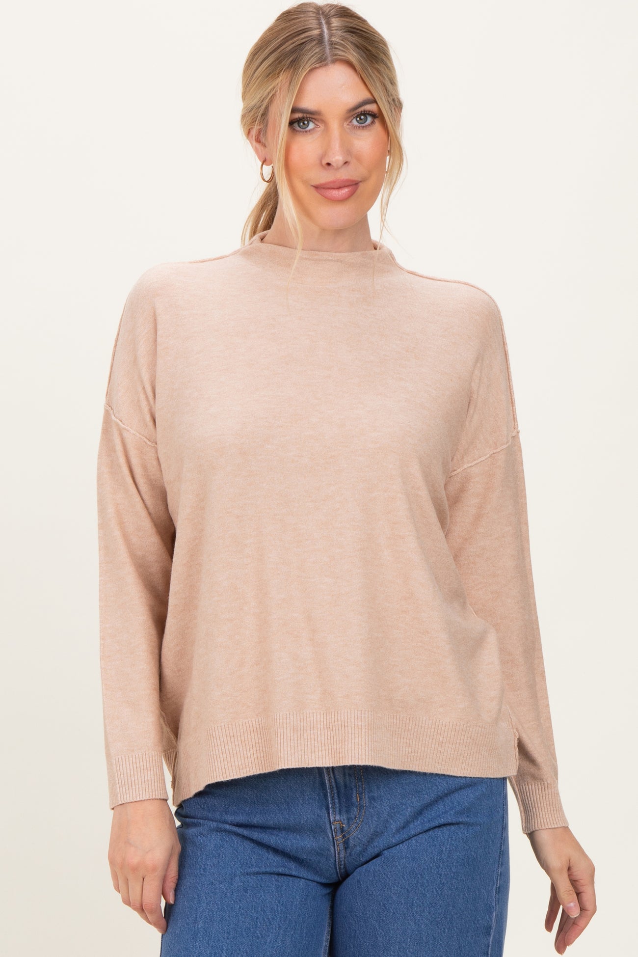 woman wearing a light beige oversized sweater with a high neckline and long sleeves, styled with blue jeans