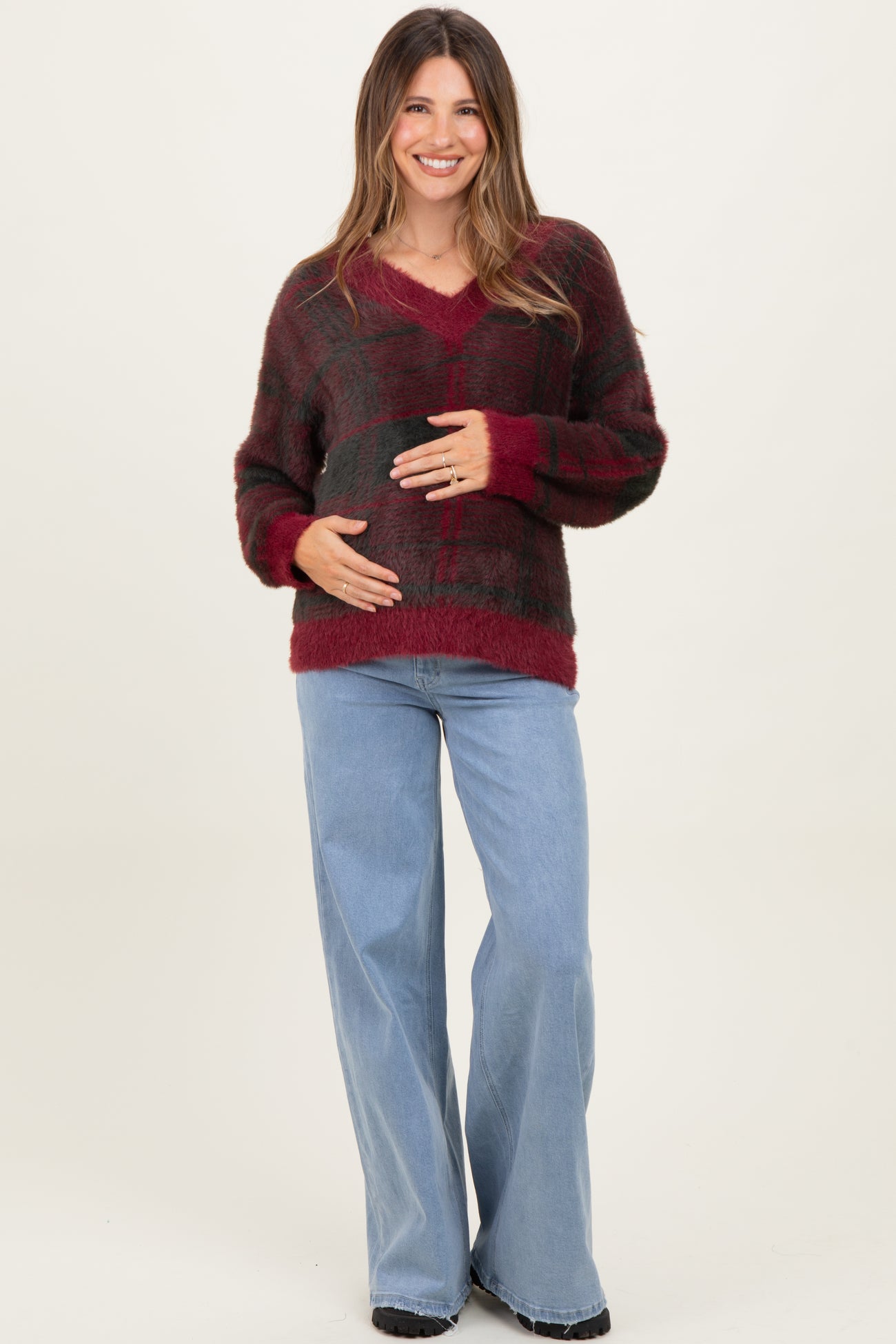 woman wearing a red and green plaid maternity sweater with a v-neck and holding her baby bump, paired with light blue wide-leg jeans