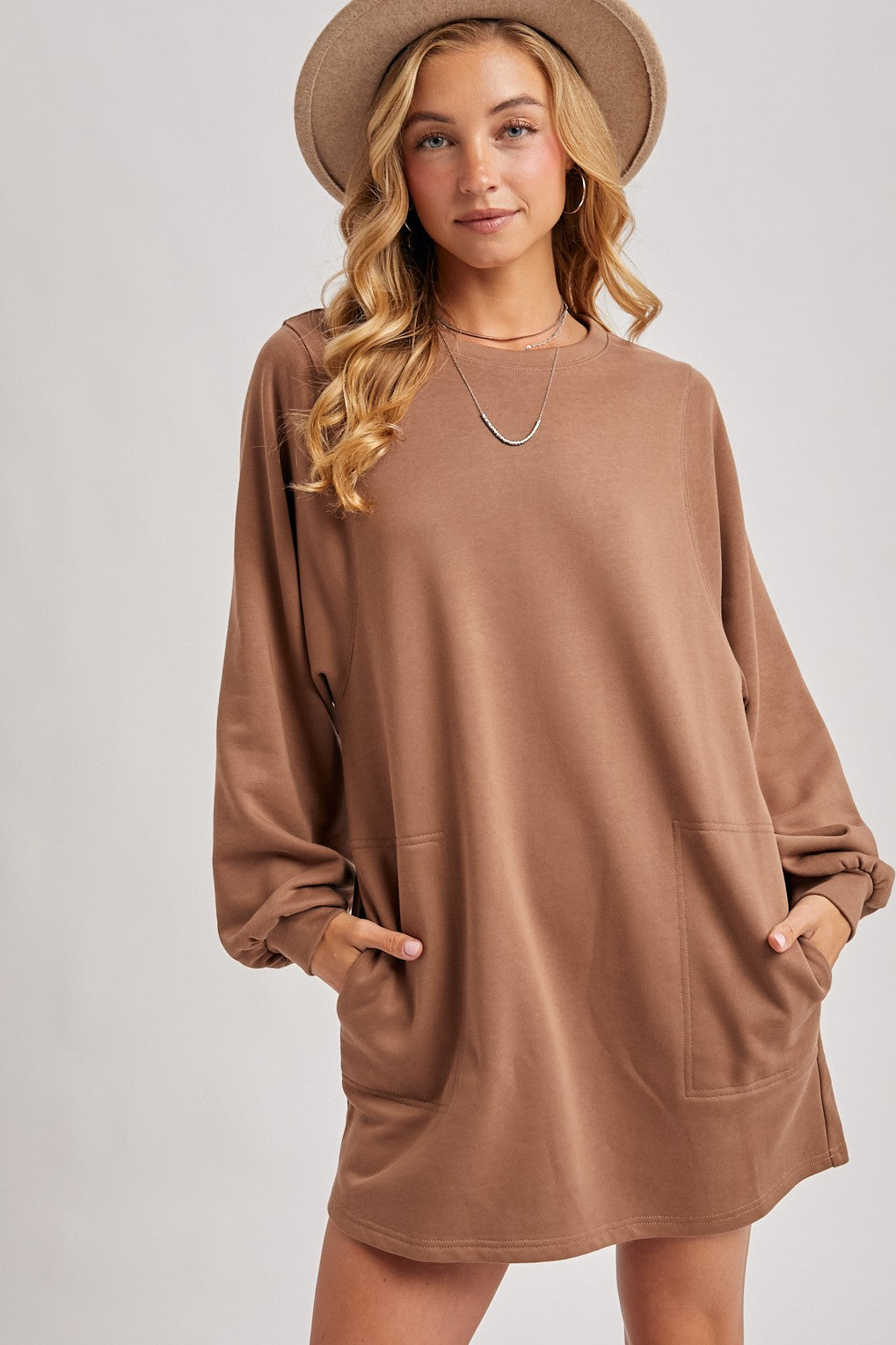 woman wearing a brown oversized sweatshirt dress with long sleeves and pockets, styled with a tan hat