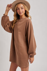 woman wearing a brown oversized sweatshirt dress with long sleeves and a front pocket, smiling while wearing a hat