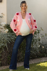 woman wearing a pink maternity cardigan with heart appliqués and blue flared jeans, showing her baby bump