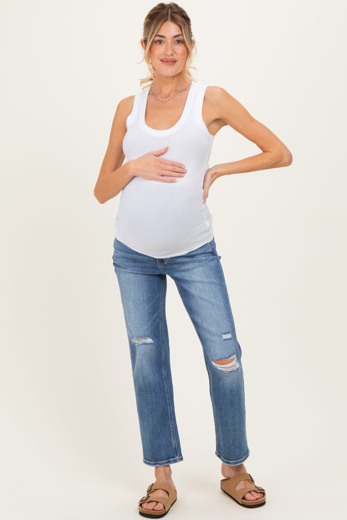 woman wearing a white ribbed maternity tank top and distressed blue maternity jeans, holding her baby bump