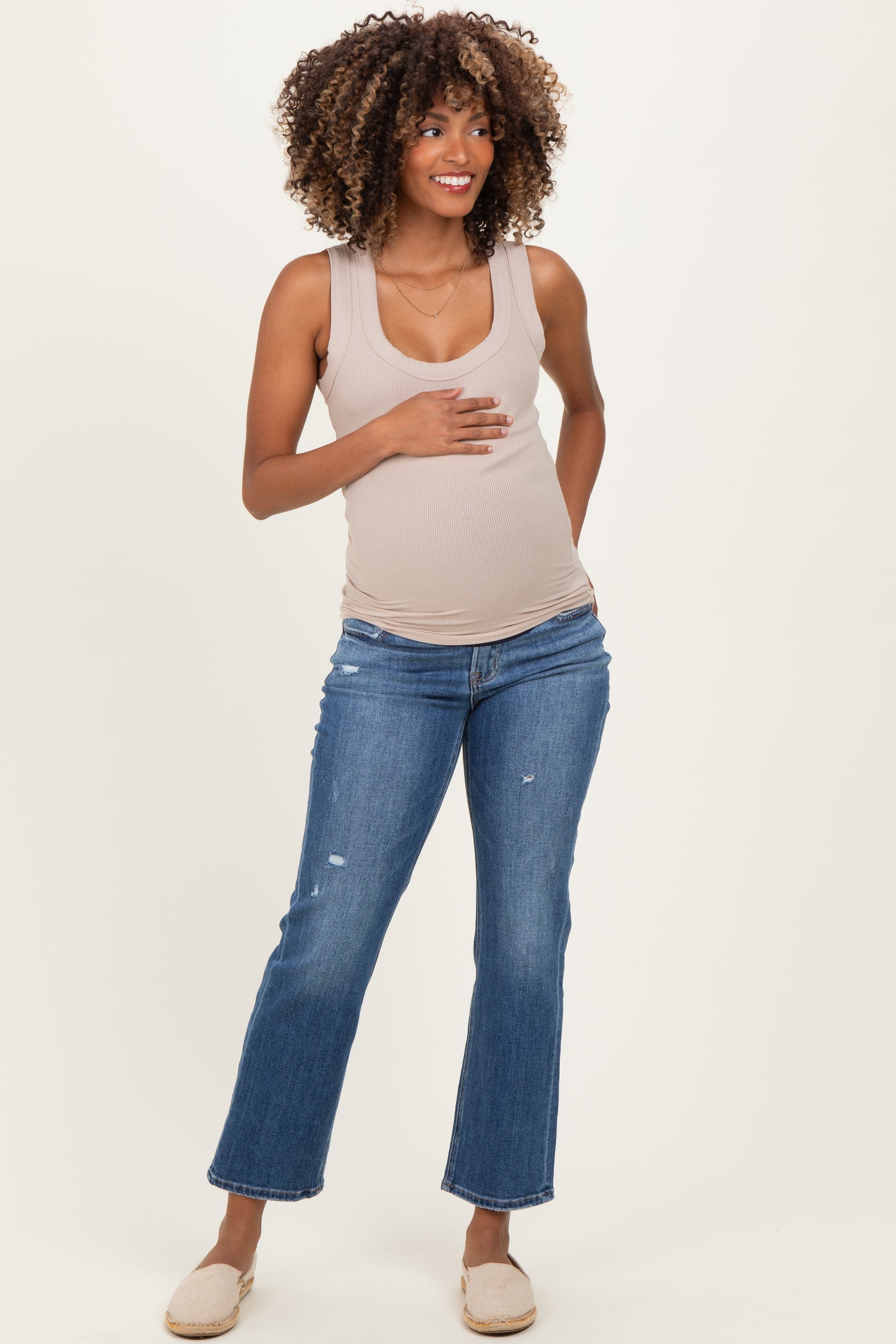 woman wearing a beige maternity tank top and distressed blue jeans, showcasing her baby bump